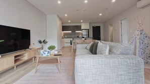 En moderne stue med et fladskærms-tv, en sofa, et sofabord og et køkken i baggrunden.