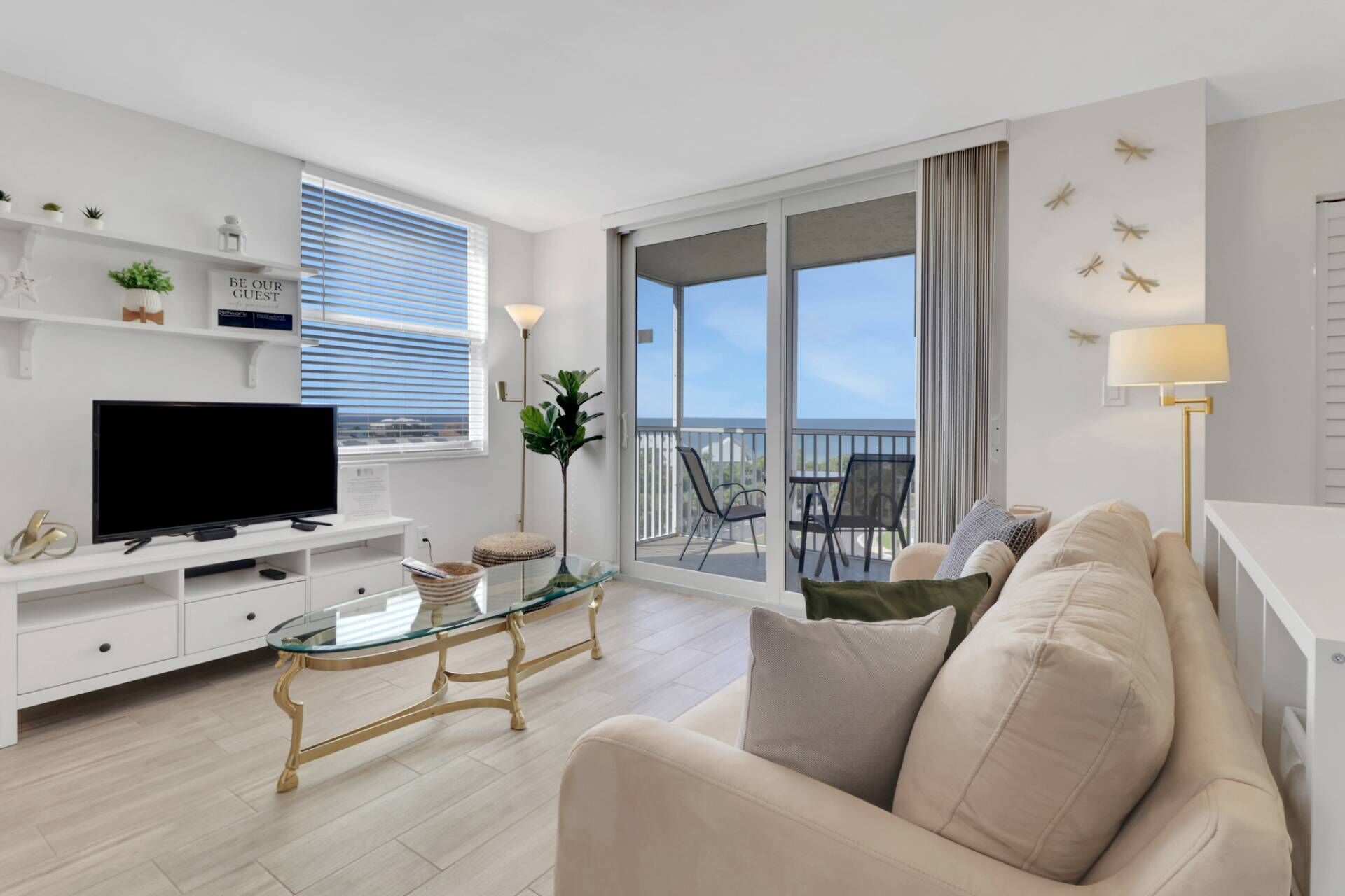 A modern living room with a flat-screen TV, a glass coffee table, a sofa, and a balcony with outdoor furniture.