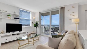 A modern living room with a flat-screen TV, a glass coffee table, a sofa, and a balcony with outdoor furniture. - GULF VIEW | 8TH FLOOR STUDIO | 2 FULL BEDS | FREE PARKING + WIFI | BEACH GEAR (Bonita Springs)