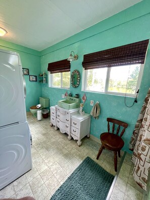 A bathroom with a washing machine, a sink, a mirror, and a window with blinds. - Cabin On The Lake (Hemphill)