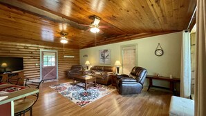 A cozy cabin interior with a wooden ceiling, a ceiling fan, a TV, a fireplace, and a dining area. - Cabin On The Lake (Hemphill)