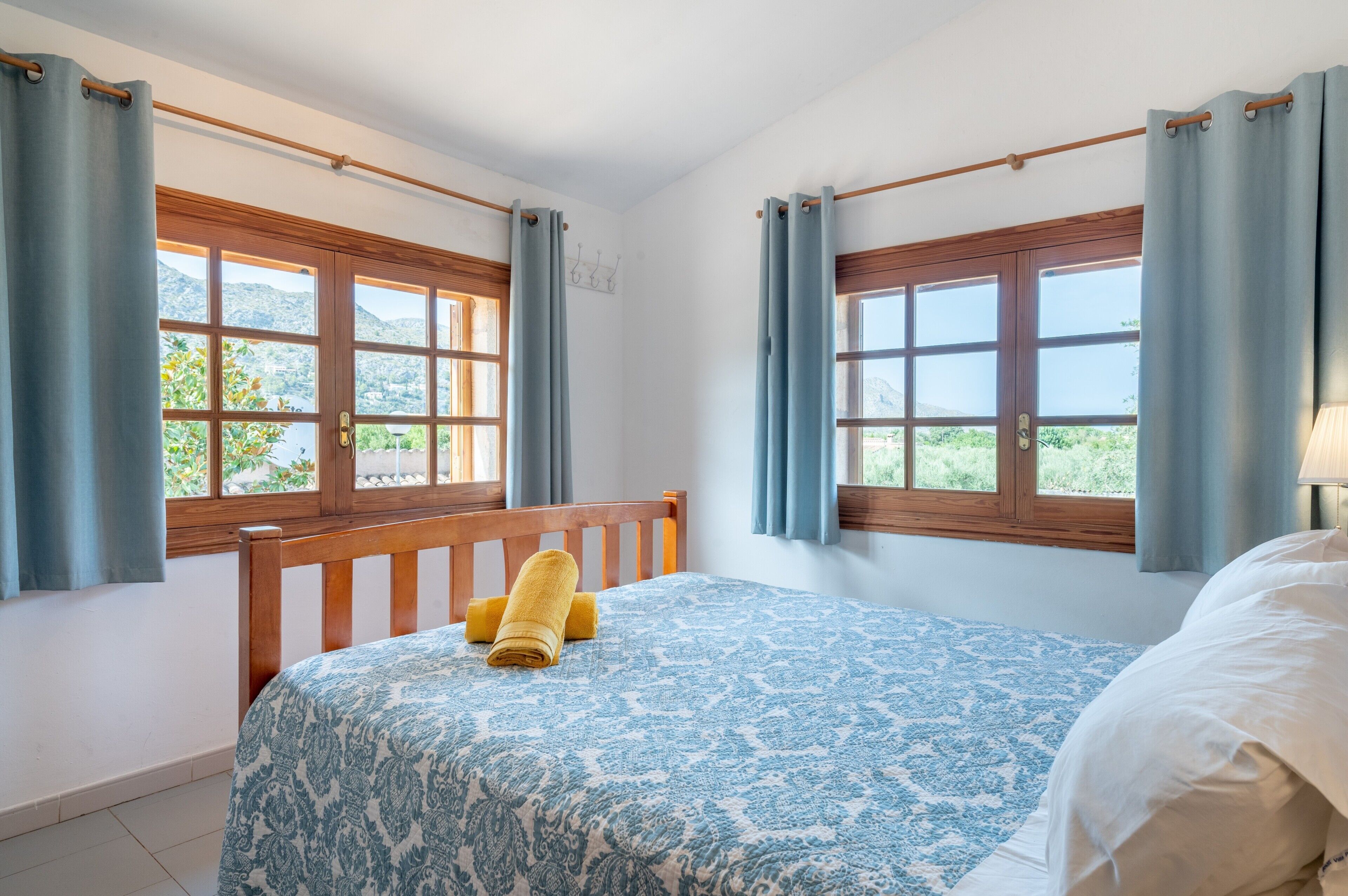 A bedroom with a bed, two windows with curtains, a wooden headboard, and a view of greenery outside.