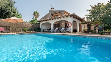 Une piscine avec des chaises longues, un bâtiment orné d’arches et un parasol en chaume.