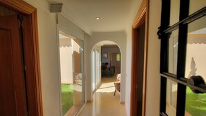 Um corredor com portas de vidro que levam a um quarto, uma porta de madeira à esquerda e vista para um jardim através de uma janela à direita.