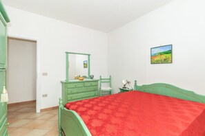 Une chambre avec un lit dont le cadre est vert, une couvre-lit rouge, une commode, un miroir, une chaise et un tableau accroché au mur.