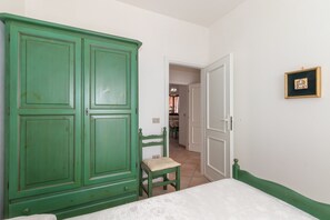 A room with a green wardrobe, a small wooden chair, a bed with a white mattress, and a framed picture on the wall.