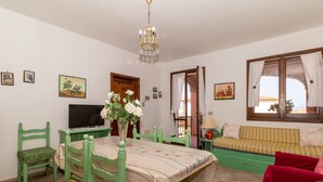 Een kamer met een eettafel, stoelen, een televisie, een bank, een raam met gordijnen en wanddecoratie.