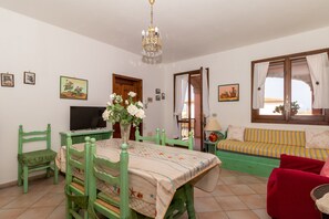 Ein Zimmer mit Esstisch, Stühlen, Fernseher, Sofa, einem Fenster mit Vorhängen und Wandbildern.