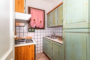 A kitchen with a gas stove, a window with pink curtains, and wooden cabinets. - Apartment  Azalea with Balcony & Wi-Fi (Valledoria)