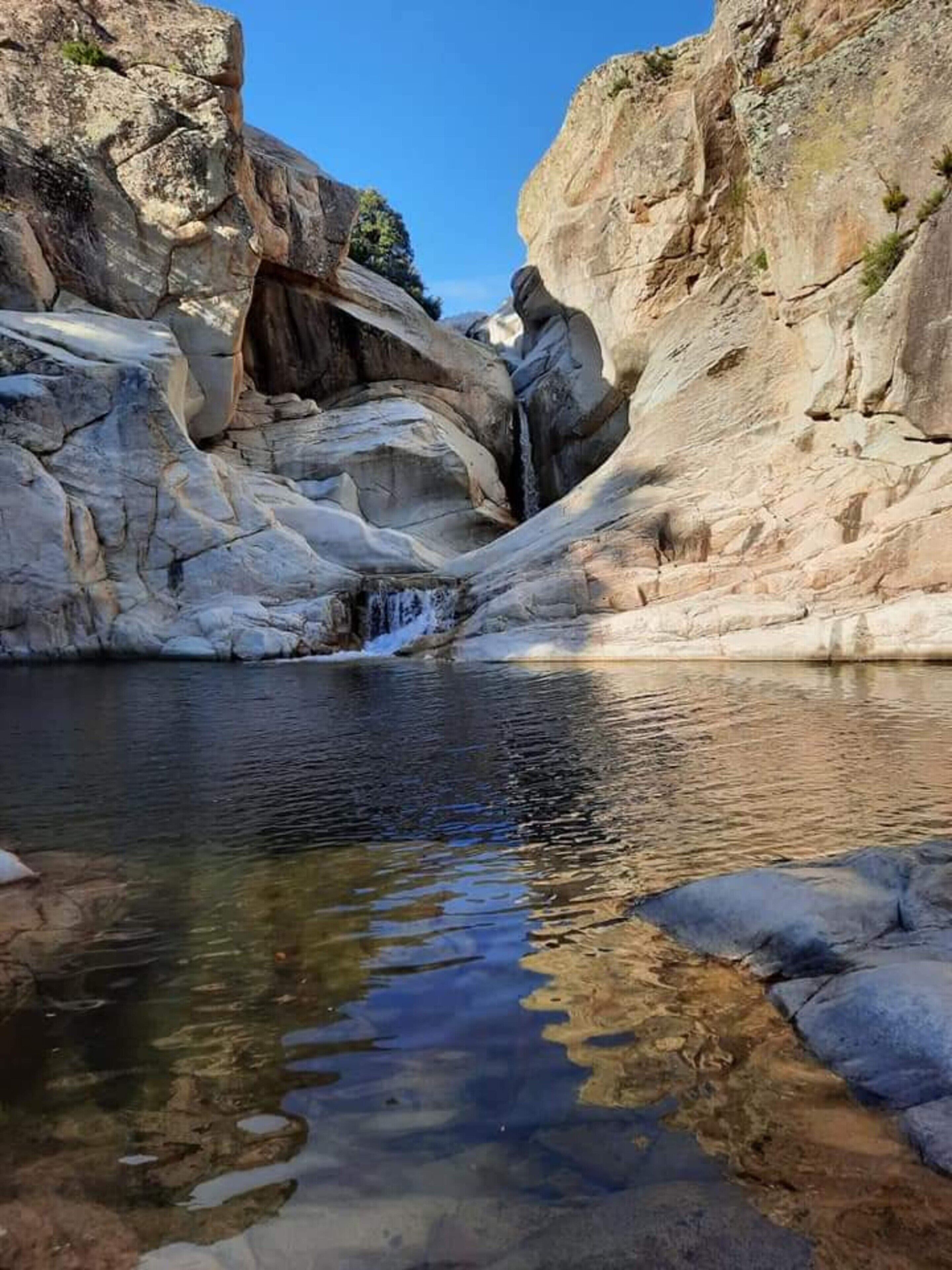 A natural rock formation with a small waterfall and clear water reflecting the surrounding rocks.