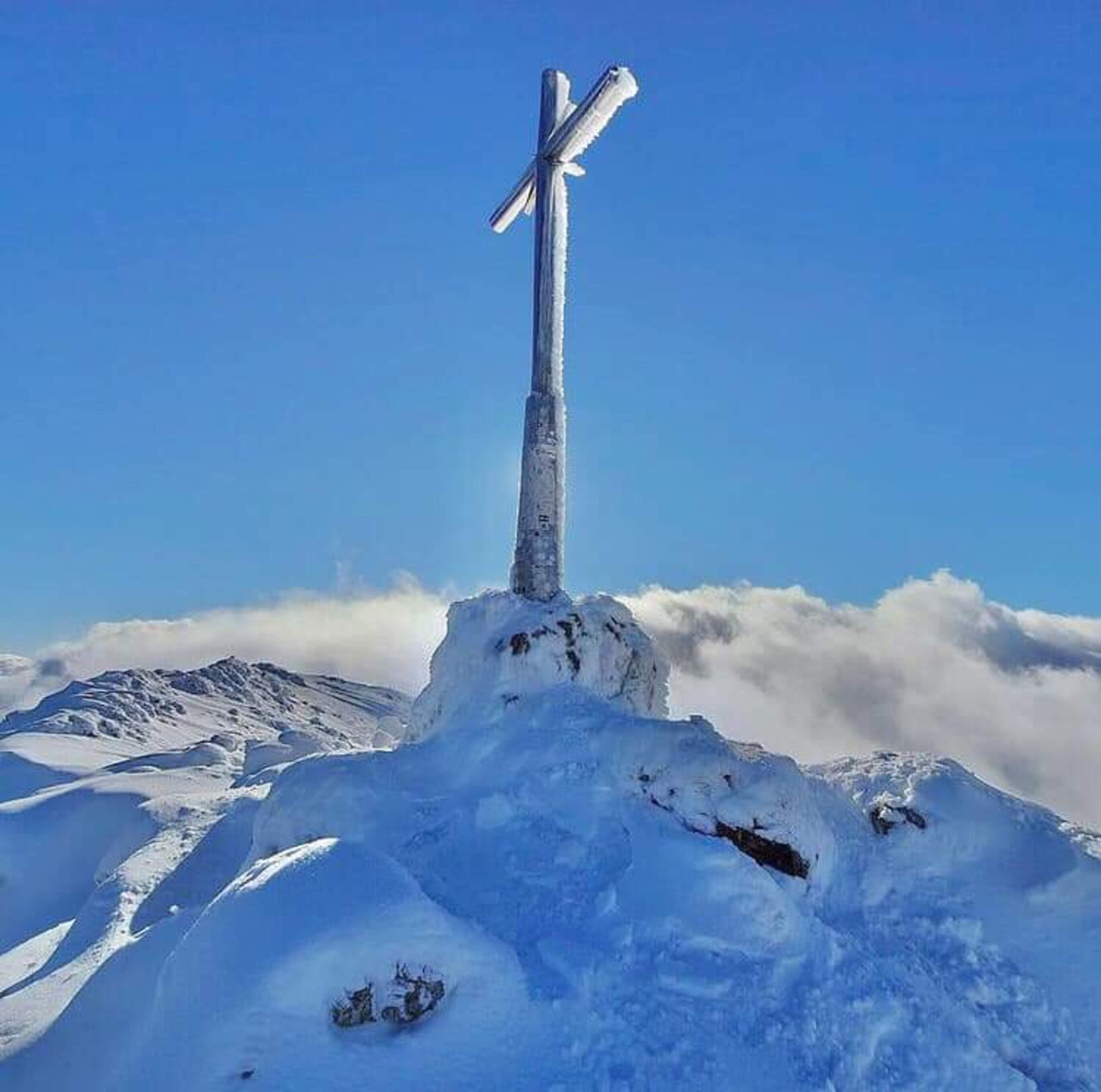 Ein schneebedecktes Kreuz auf einem Berggipfel bei klarblauem Himmel.