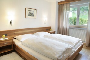 A double bed with white linens, two bedside tables with lamps, a framed picture on the wall, and a window with curtains.