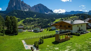 A mountain village with a prominent peak in the background, wooden houses, and a paved area with a pool.