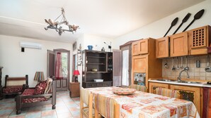 A room with a bed, a dining table with a tablecloth, a kitchen area with wooden cabinets, and a chandelier. - Holiday Home "Sa Burrida" with Garden, Covered Terrace & Fireplace (posada)