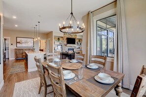 Un coin repas avec une table en bois dressée pour quatre personnes, entourée de chaises. Un lustre est suspendu au-dessus, et une cheminée en pierre est visible au fond.