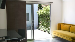 Une pièce dotée d’une porte-fenêtre coulissante donnant sur un jardin, d’un canapé jaune, d’une table avec une chaise et d’un téléviseur à écran plat fixé au mur.
