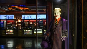 Un mannequin vêtu d’un costume violet avec une cravate rayée, tenant un chapeau violet, se tient devant l’enseigne d’un cinéma TCL Chinese IMAX.