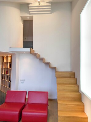 Una escalera con peldaños de madera que conduce a un piso superior, una zona de estar roja y una estantería con libros.