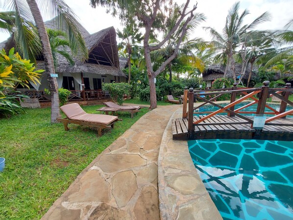 A pool area with lounge chairs, a wooden deck, and a thatched-roof building.