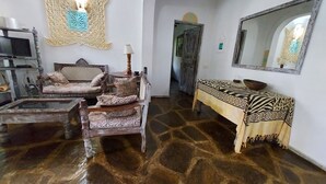 Una sala de estar con piso de piedra, un conjunto de sofás, una mesa con mantel estampado, un espejo y un adorno de pared.
