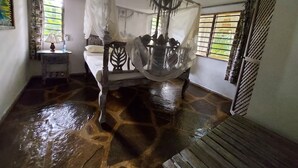 Una habitación con cama bajo dosel, una mesita de noche con lámpara, una ventana con vistas y un ventilador de techo.