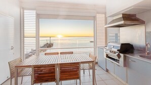 Un balcon offrant une vue sur le coucher de soleil sur la mer, une table et des chaises en bois, et un barbecue encastré.