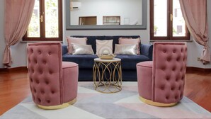 Een kamer met een donkerblauwe bank, roze pluchen ottomaantjes, een bijzettafeltje met gouden rand en een grote spiegel boven de bank.