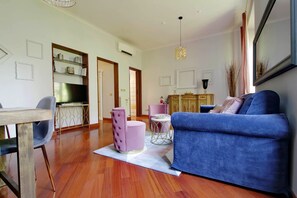 Un salon moderne avec un canapé bleu, une table à manger avec des chaises, un pouf rose et un coin cuisine équipé d’un téléviseur.