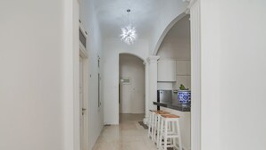 Un pasillo con paredes blancas, suelo de baldosas y una lámpara en el techo. Se observan columnas blancas y una puerta al final del pasillo.