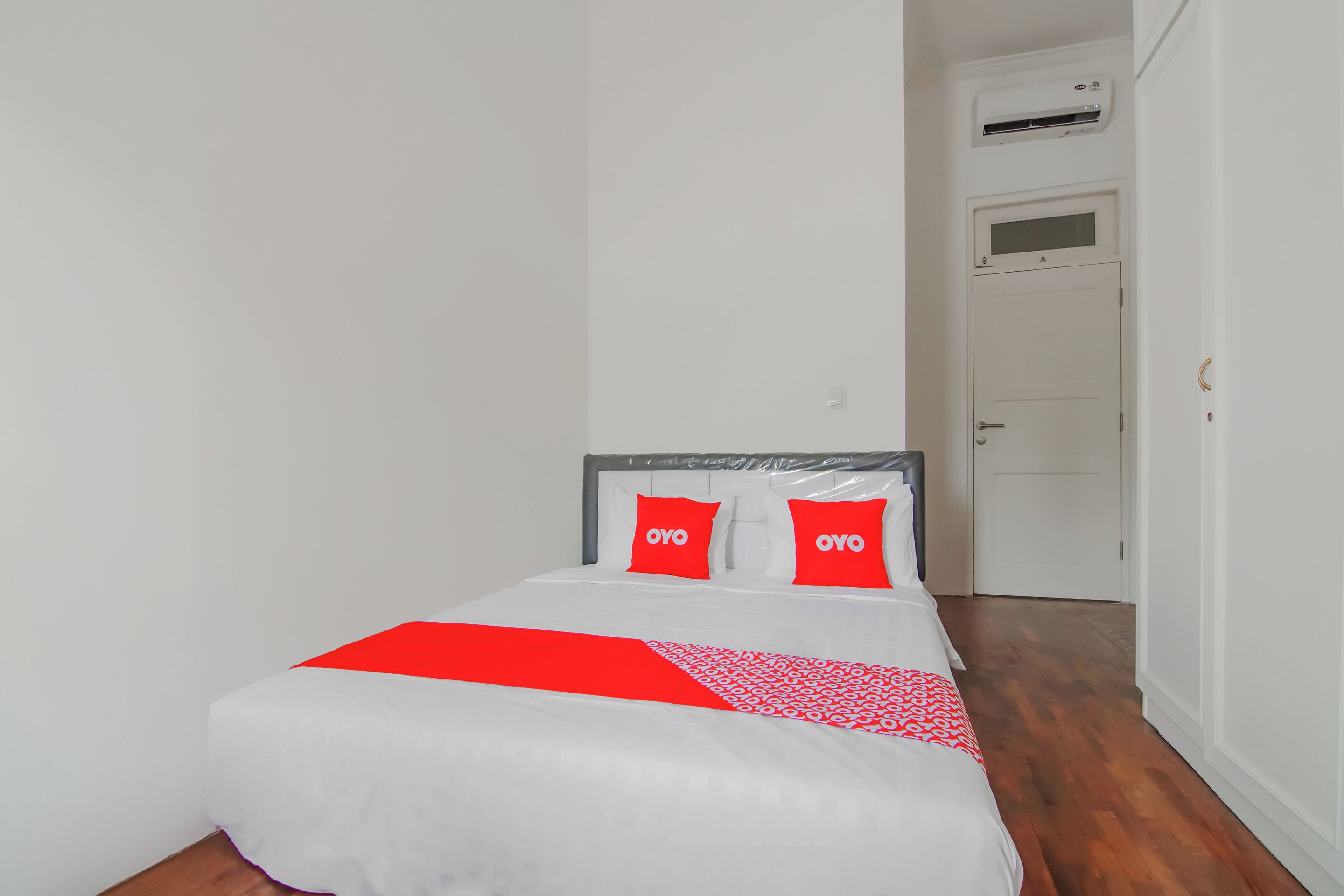 A neatly made bed with white and red bedding, a grey headboard, and two red pillows with the word "OYO" on them.