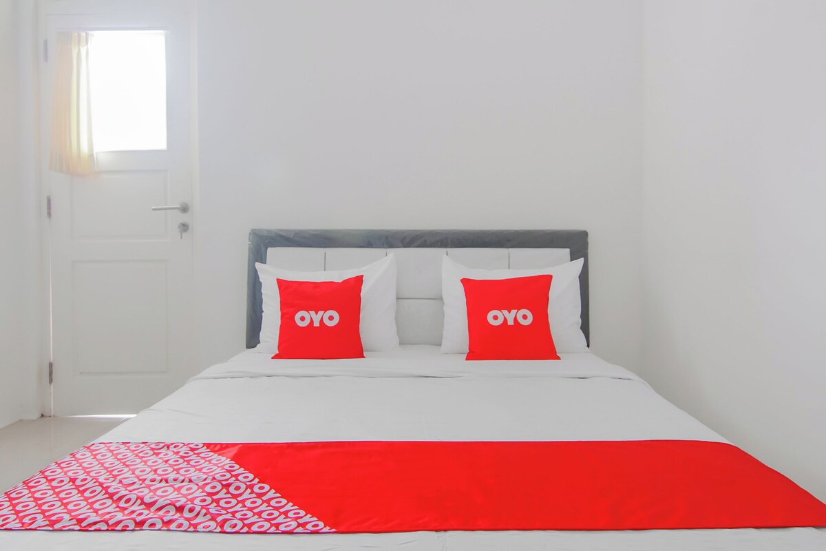 Un lit avec une literie blanche et deux oreillers rouges ornés du texte « OYO », une porte blanche et une fenêtre avec des rideaux.
