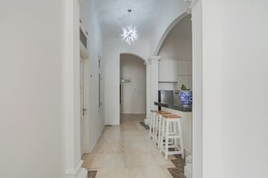 Un couloir aux murs blancs, doté d’un sol carrelé et d’un luminaire au plafond. On y trouve des colonnes blanches et une porte au fond du couloir.