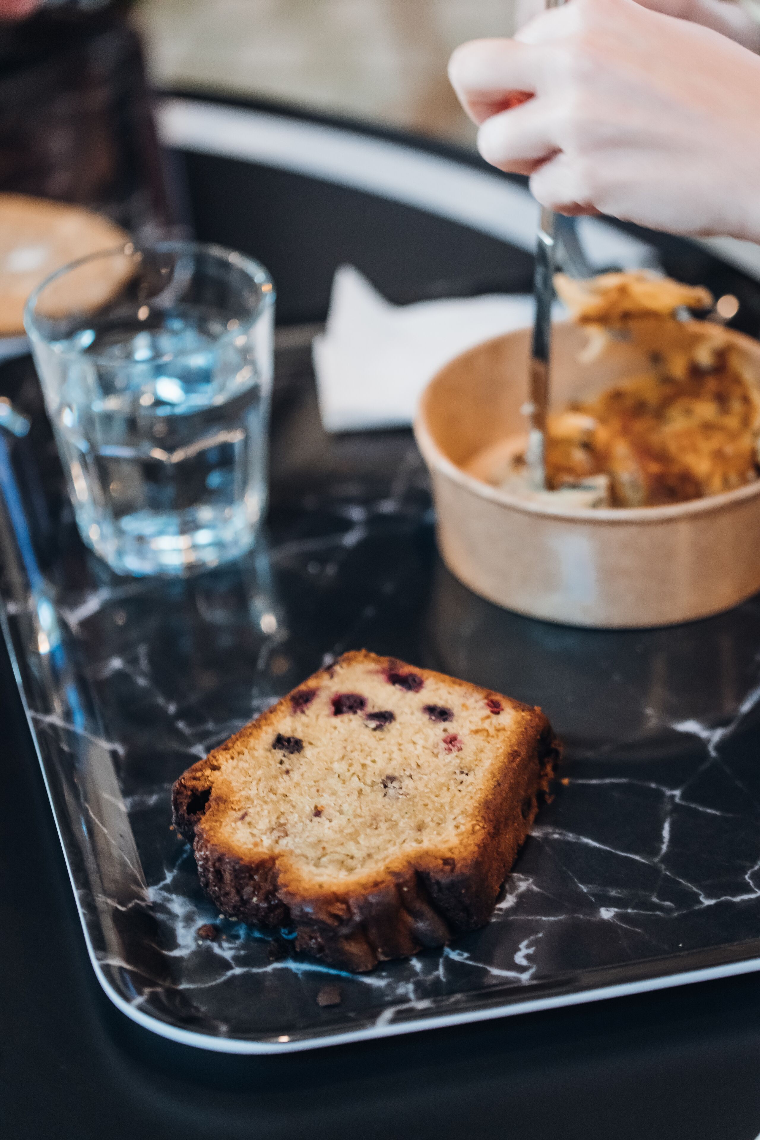 Une part de gâteau sur un plateau en marbre, accompagnée d’un verre d’eau et, au fond, d’un bol rempli de gâteau.
