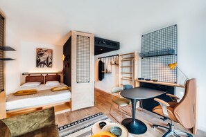 A compact, modern room with a bed, a desk, and a chair. There is a small table with a lamp, a shelf with storage boxes, and a wall-mounted rack.