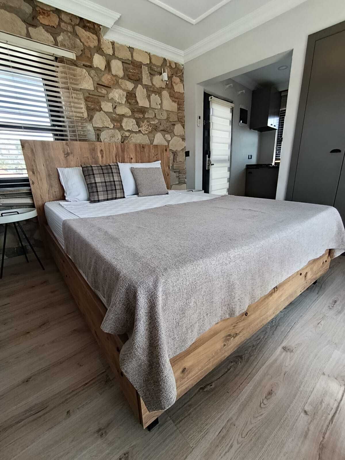 A bedroom with a stone wall, a wooden headboard, a bed with a grey blanket, a small table, and a door leading to a kitchen.