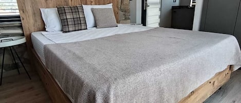 A bedroom with a stone wall, a wooden headboard, a bed with a grey blanket, a small table, and a door leading to a kitchen.