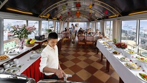 A dining car with tables set for a meal, a waiter serving food, and a view of the city through large windows. - Hanoi Posh Hotel (Hanoi)