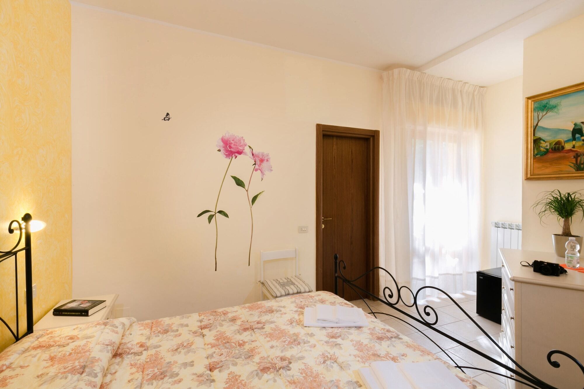 A bedroom with a bed, a door, a window with curtains, a wall-mounted lamp, and a painting.