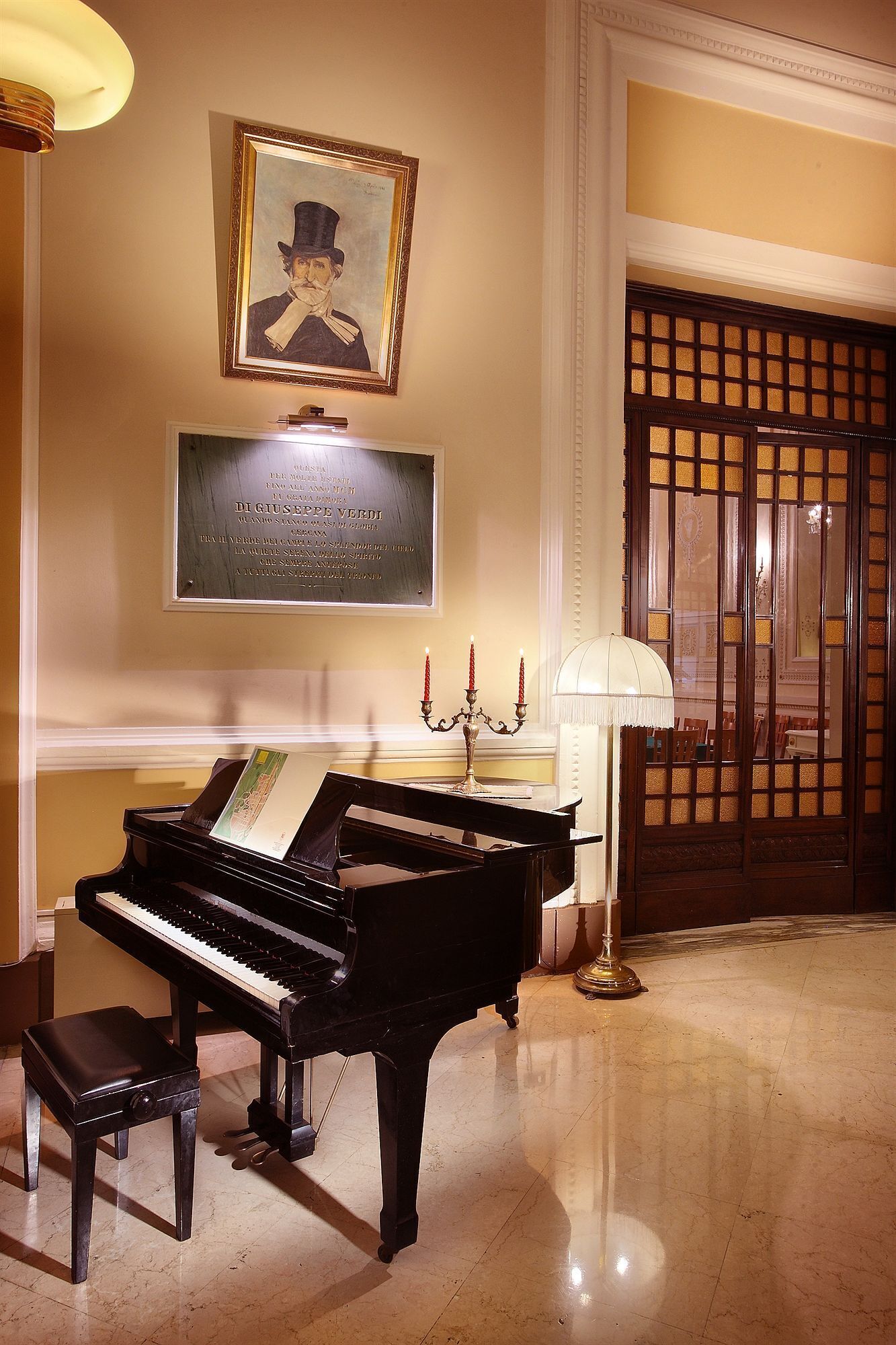 Un piano à queue dans une pièce bien éclairée, avec un portrait encadré au-dessus, un chandelier avec des bougies et une porte en bois traditionnelle.