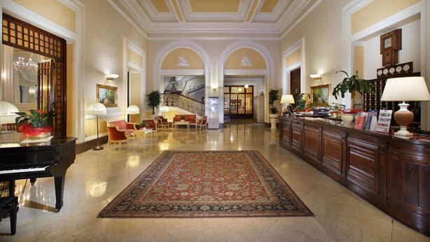 A spacious lobby with a grand piano, a large ornate rug, and a reception desk with a plant.