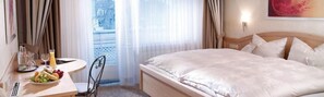 A hotel room with a bed, a bedside table with a fruit plate, a chair, and a window with curtains. - Hotel Willinger Hof (Willingen)