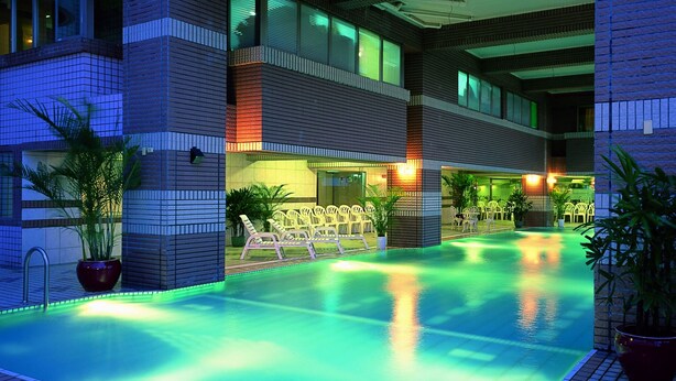 A swimming pool with a tiled edge, surrounded by lounge chairs and potted plants, with a building featuring a modern design in the background. - Cheng Pao Hotel (Puli)
