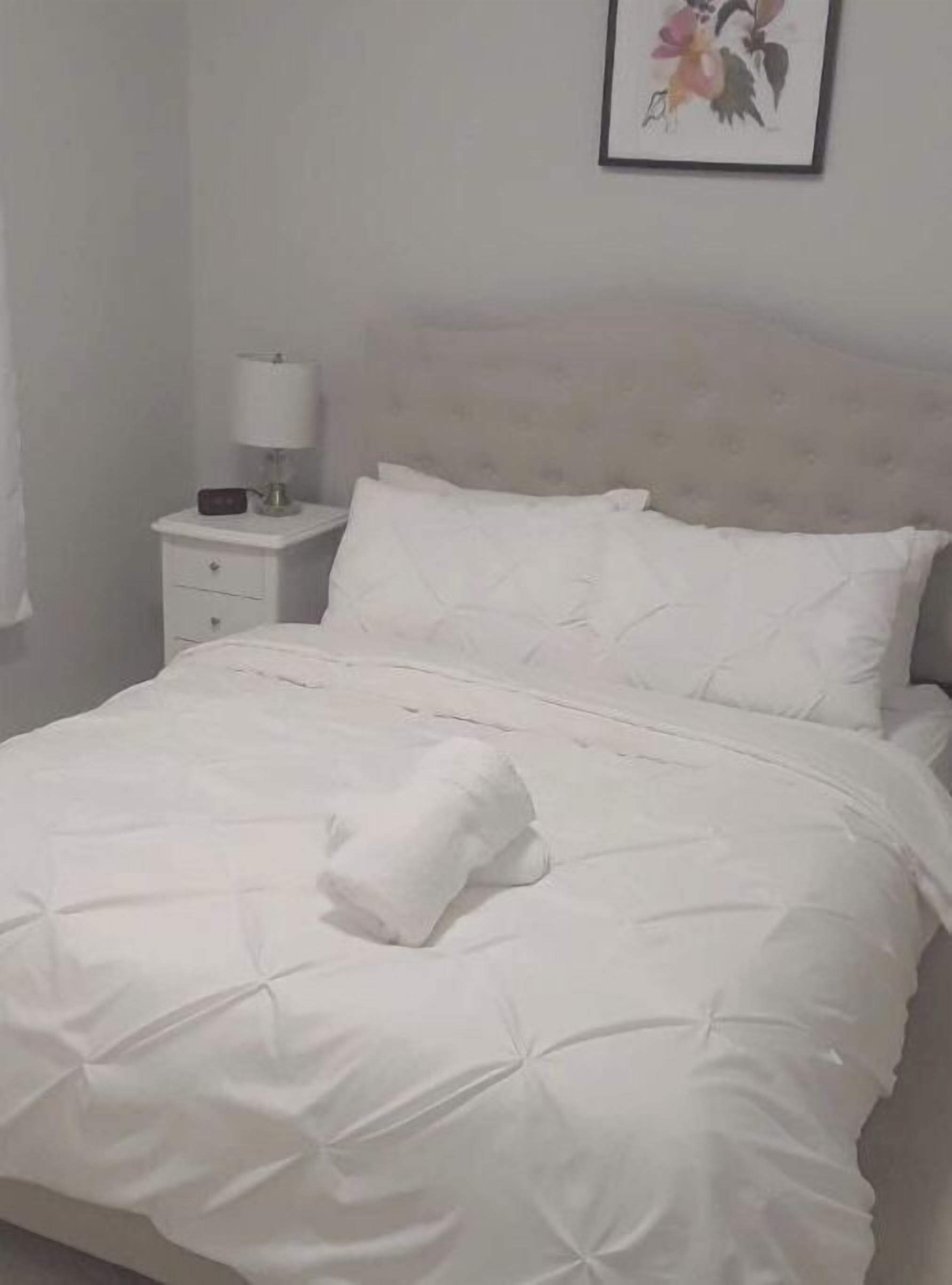 Una cama bien hecha con edredón blanco y almohadas, una mesita de noche con lámpara y un cuadro en la pared.