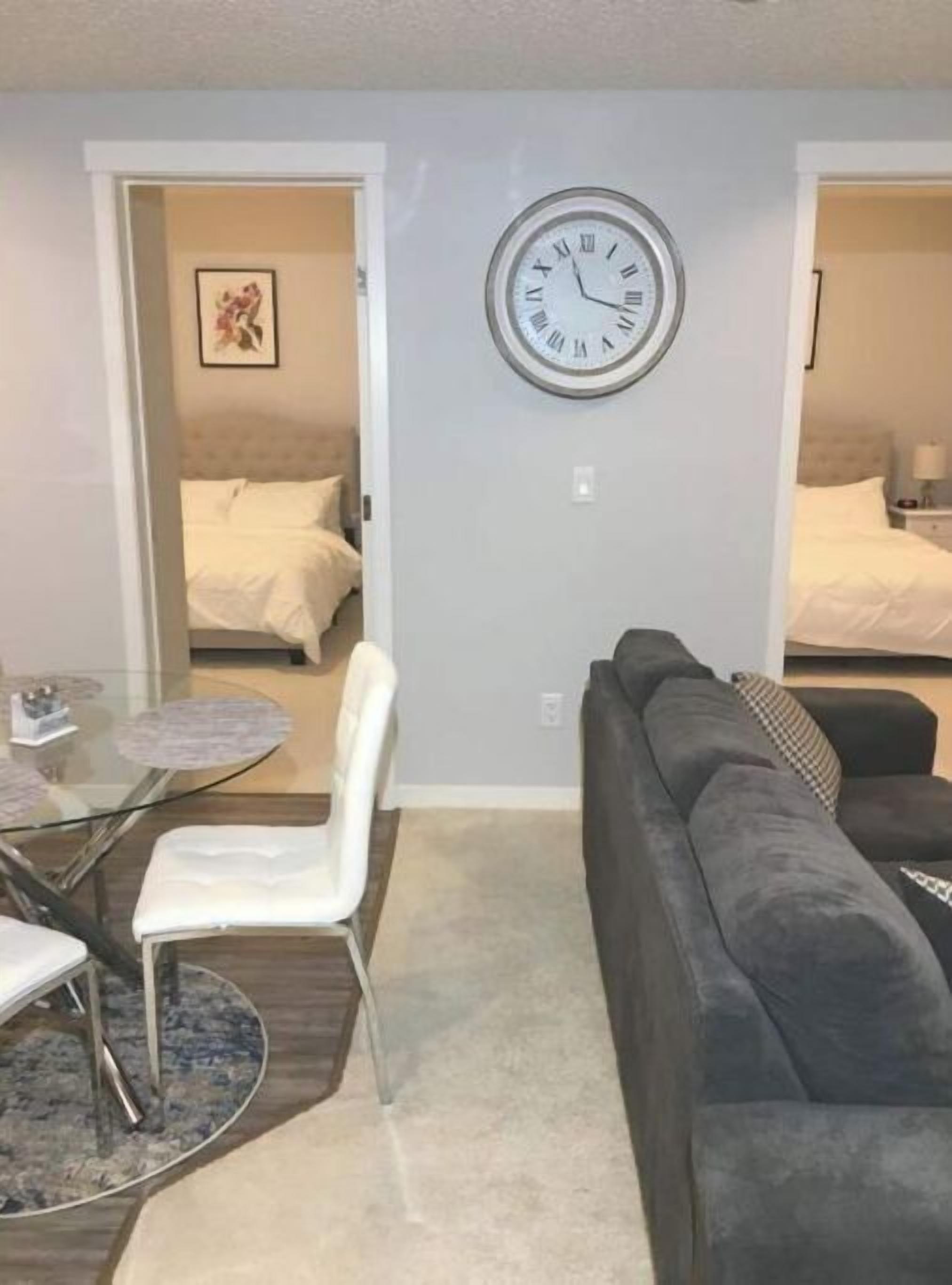 Ein modernes Wohnzimmer mit grauem Sofa, einem Esstisch mit Stühlen und einer großen Wanduhr.