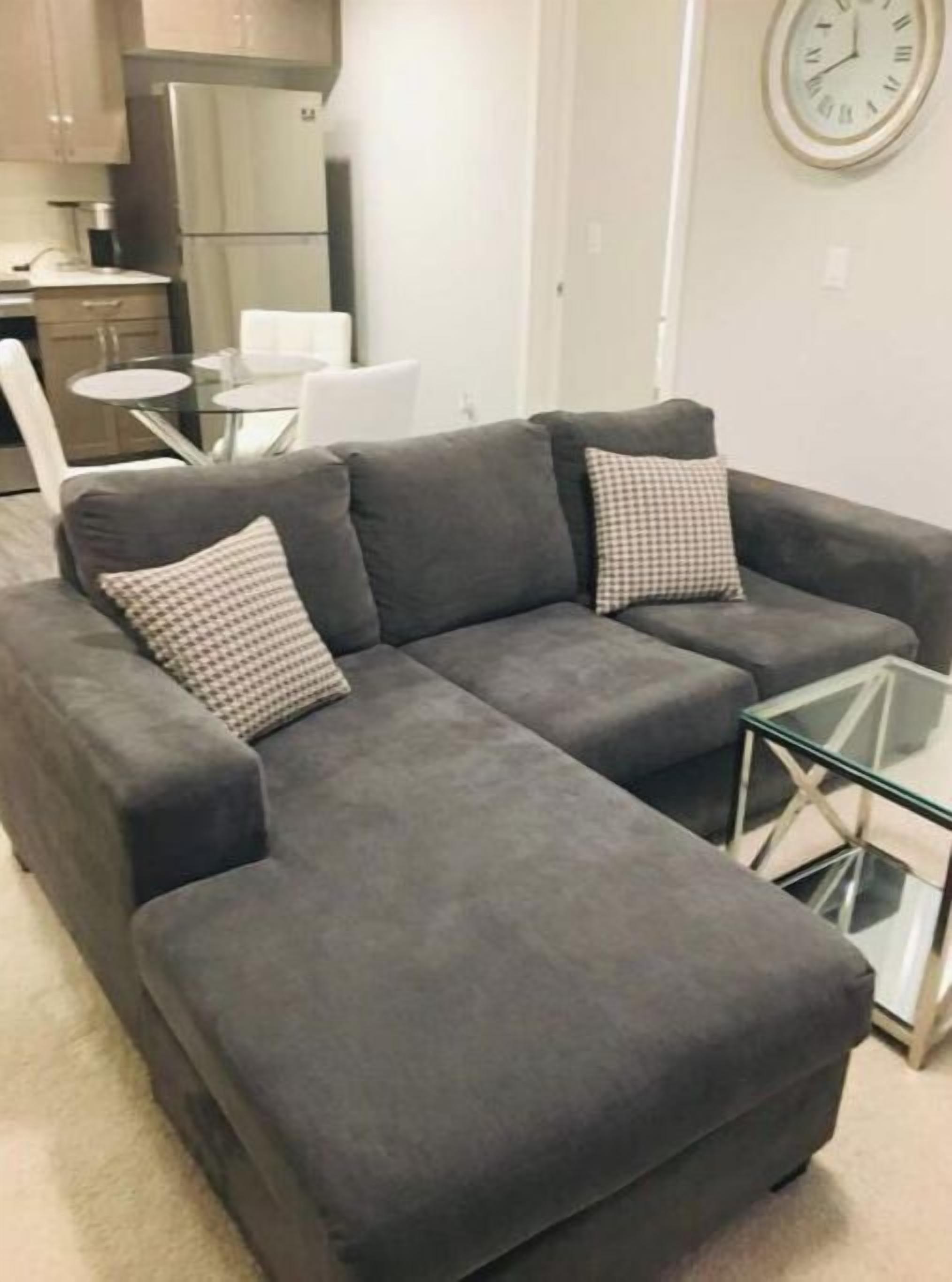 Ein graues Ecksofa mit karierten Kissen, einem Glastisch und einer Wanduhr.