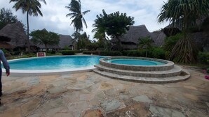 A swimming pool with stone steps leading into it, surrounded by palm trees and thatched-roof structures. - Harbour Key Cottages (Malindi)