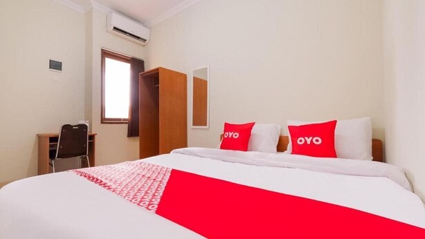 A hotel room with a bed, two red pillows with "OYO" branding, a wooden wardrobe, a desk with a chair, and a window.