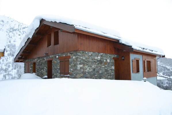 A wooden cabin with stone walls and wooden shutters, surrounded by snow.