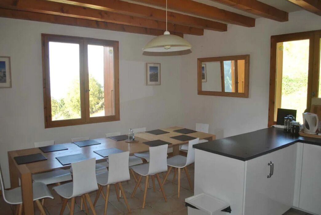 A dining area with a wooden table and chairs, a kitchen counter, and large windows with wooden frames.