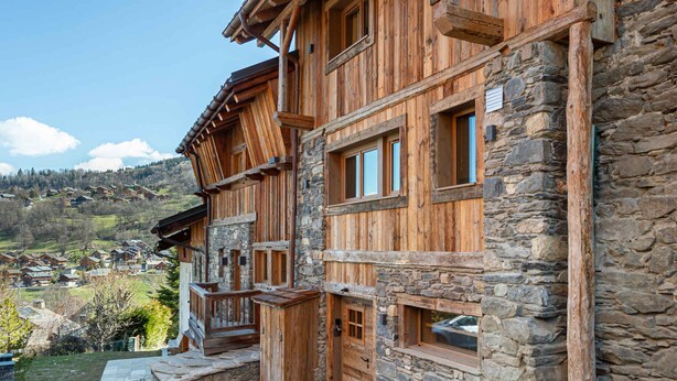 A wooden chalet with stone walls, a balcony, and a view of a village in the background. - Cozy Chalet with Garden, Near Ski Lifts and Free Bus (Méribel)